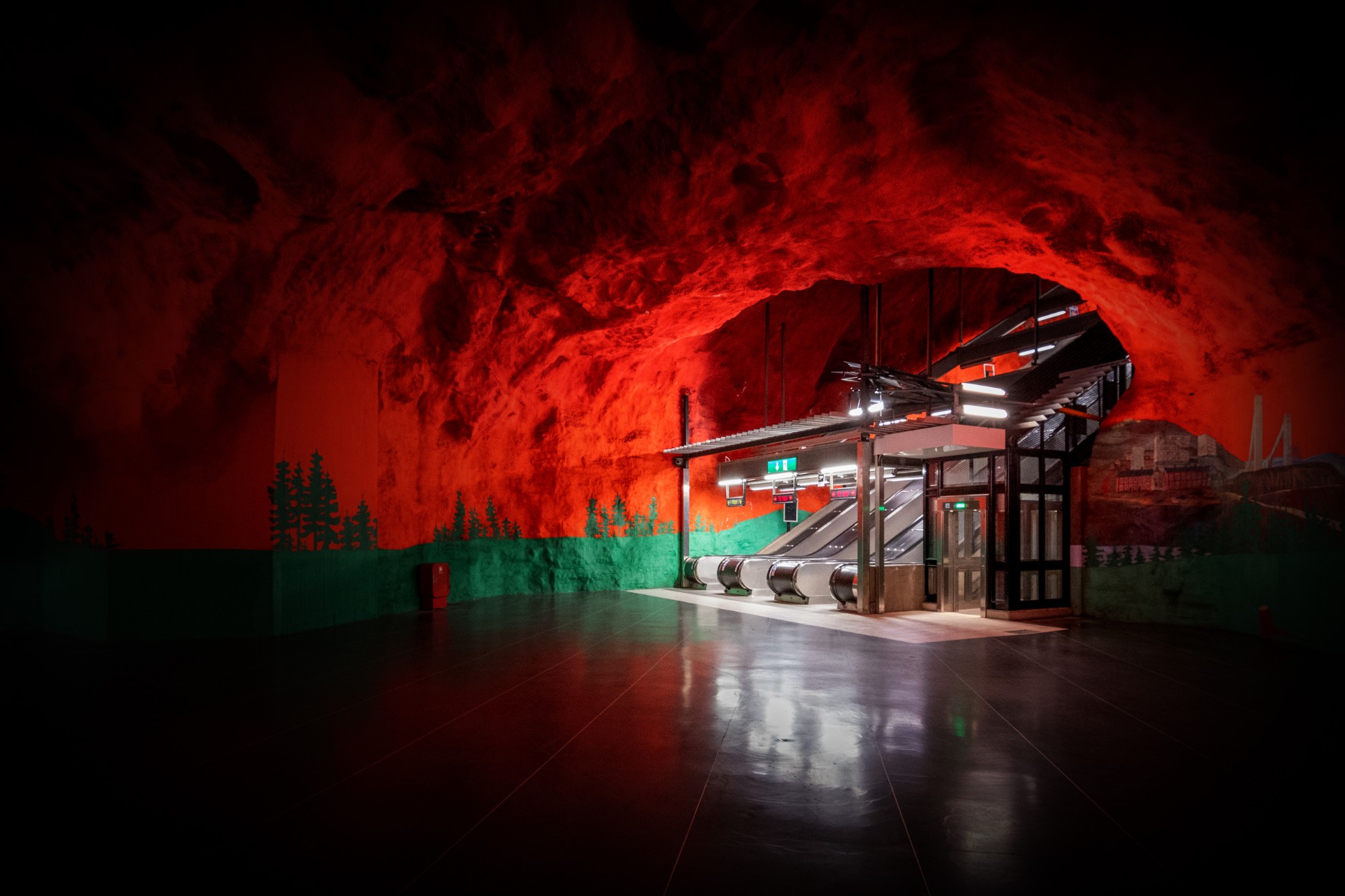 Station de métro creusée dans la roche éclairée en rouge, avec une fresque verte de forêt et des escalators