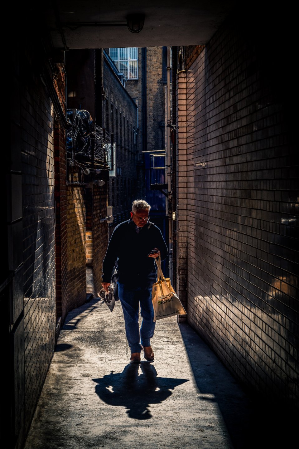 Homme qui marche en consultant son téléphone dans un passage étroit en briques, lumière rasante, ombre portée au sol et sac en toile à la main