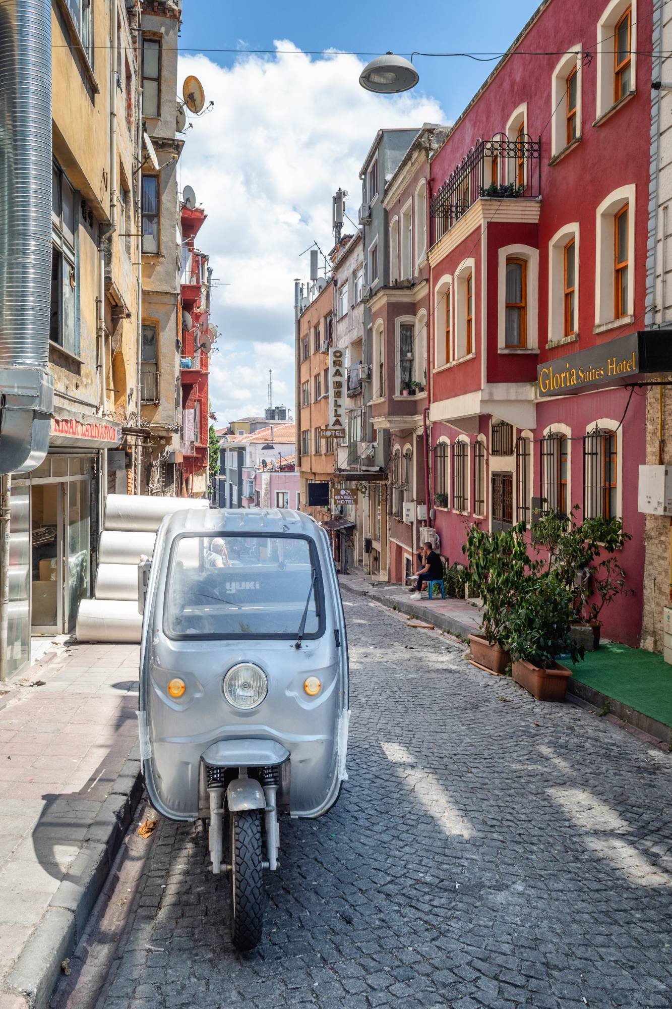 Rue pavée en pente bordée de façades colorées, avec un petit véhicule utilitaire à trois roues argenté au premier plan