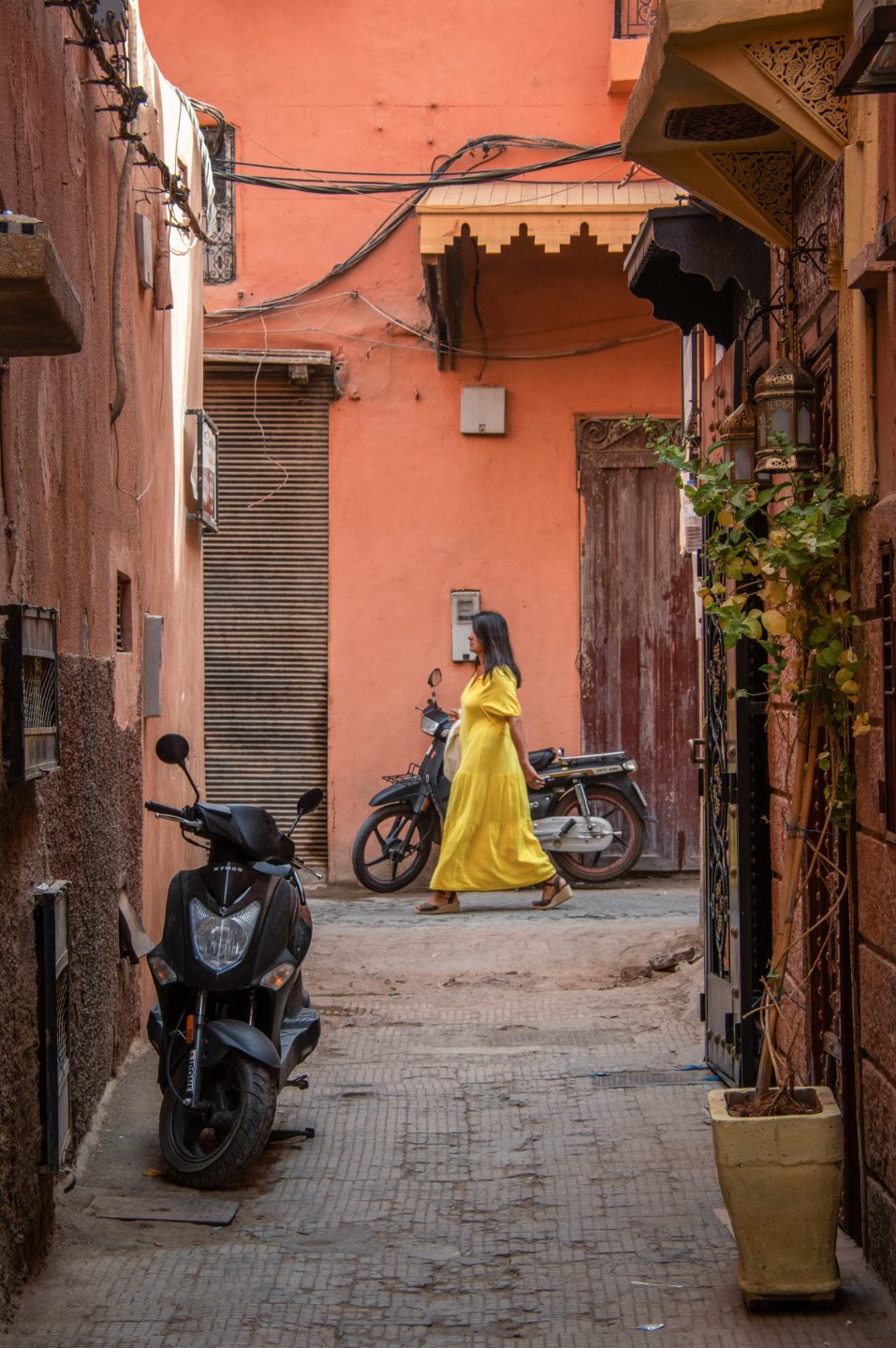 Ruelle aux murs orange où une femme en robe jaune passe devant un scooter et une porte en bois, lanternes ouvragées sur la droite