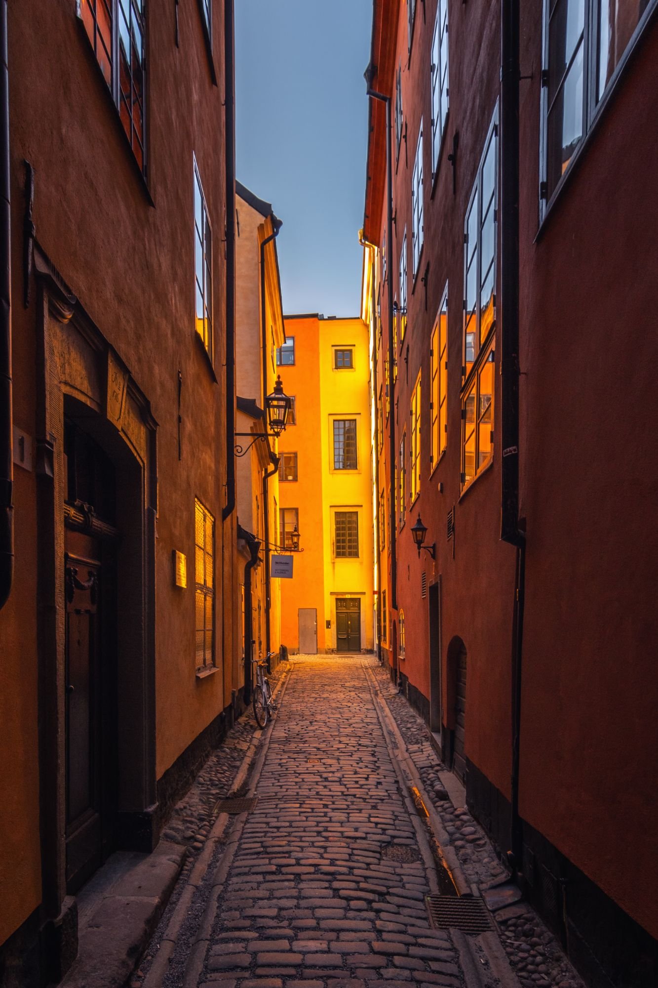 Ruelle pavée au crépuscule entre des façades orange et rouges, avec un immeuble jaune éclairé au fond et un vélo contre le mur